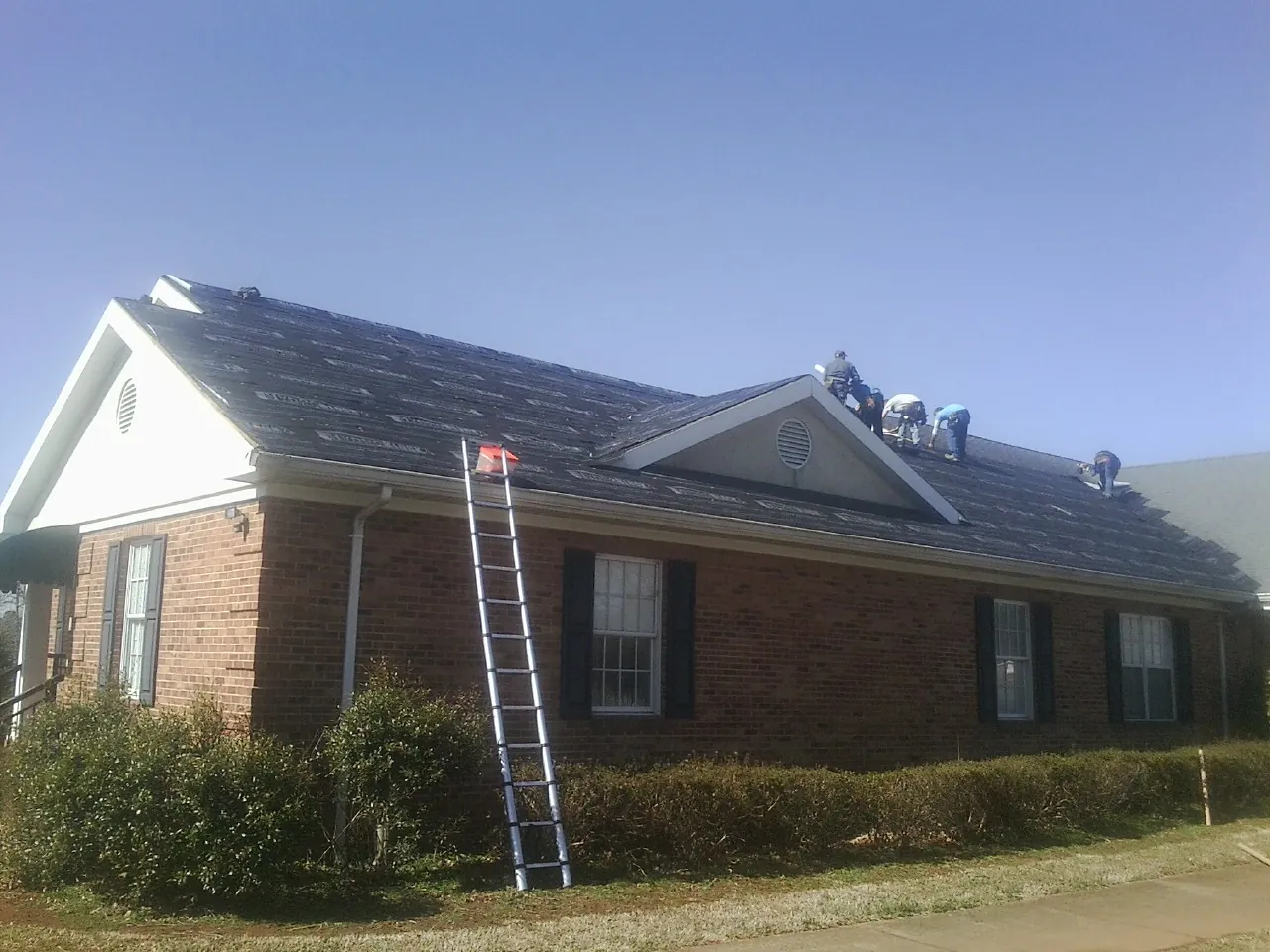 Crew tearing shingles off a brick ranch roof with a ladder set — Metal Roof Repair work by our crew in Goehner