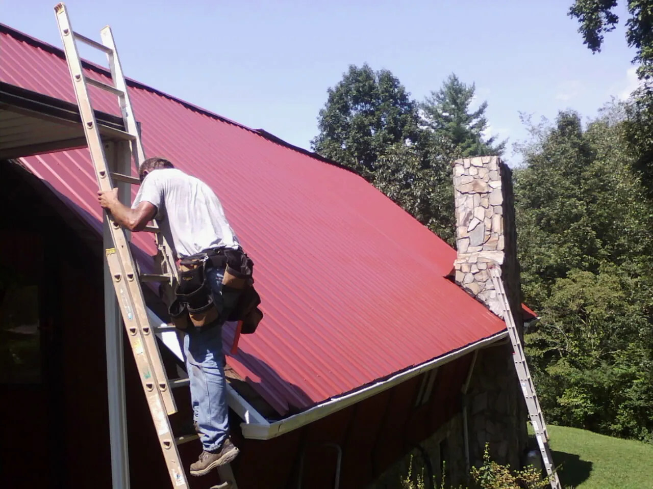 Roofer on a ladder beside a red metal roof and stone chimney — Soffit & Fascia Repair by Roofing Brothers in Goehner